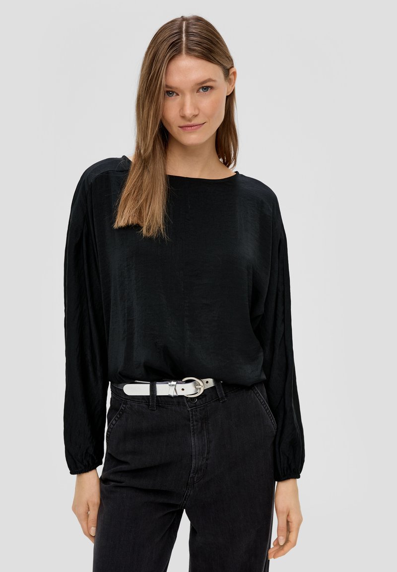 Schwarze Bluse mit langen Ärmeln, glatter Textur, runder Ausschnitt und lockerer Passform, kombiniert mit dunklen Jeans und einem weißen Gürtel mit einer runden Schnalle.