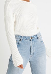 Valkoinen neulepaita, jossa on rento istuvuus ja leveät hihat, yhdistettynä vaaleansinisiin korkeavyötäröisiin farkkuihin. Ei merkittäviä kuvioita tai yksityiskohtia.