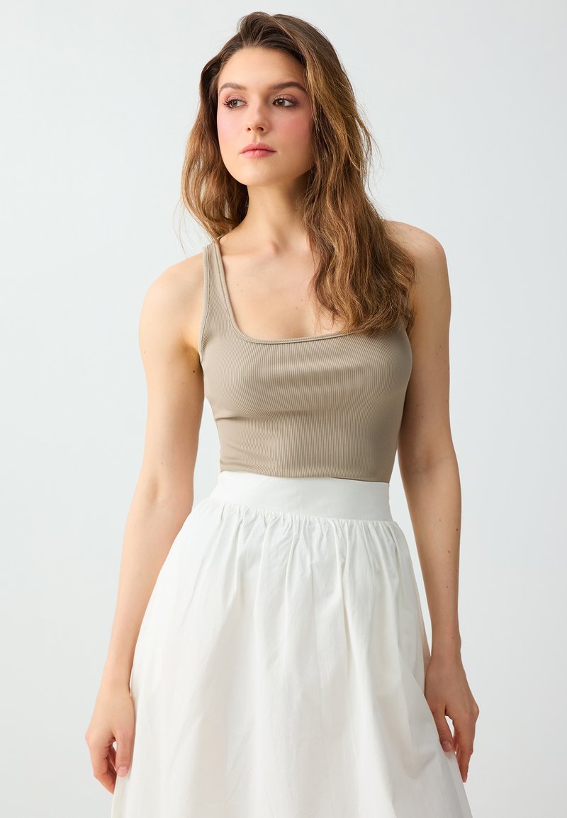 Jeune femme aux longs cheveux ondulés portant un haut beige sans manches et une jupe blanche taille haute, regardant vers sa gauche sur un fond uni.