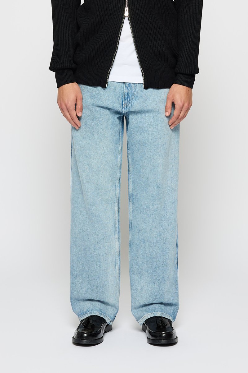 Persoon in lichtblauwe wijdvallende jeans, zwarte leren tassellaarzen, een wit shirt en een zwarte zip-up trui.