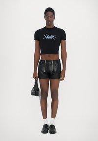 Schwarzes, verkürztes T-Shirt mit "GmbH" Grafikdesign, kombiniert mit schwarzen Leder-Shorts mit Reißverschlüssen. Das Modell hält eine strukturierte schwarze Tasche.