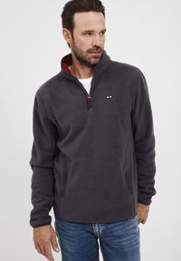 Pelēks flīsa džemperis ar ceturtdaļas rāvējslēdzēju, sarkanu iekšējo apkakli, sānu kabatām un mazu logo uz krūtīm. Mīksta struktūra, parasts piegriezums.