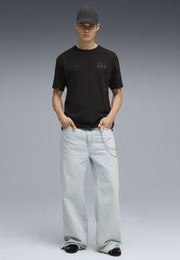 Svart t-shirt med korta ärmar som har grafiska detaljer, kombinerad med ljusblå vida jeans och en svart keps, kompletterad med en kedjedetalj.