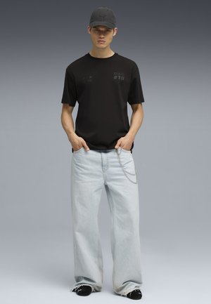 Camiseta negra de manga corta con acentos gráficos, combinada con pantalones vaqueros de pierna ancha en un tono azul claro y una gorra negra, complementada con un detalle de cadena.