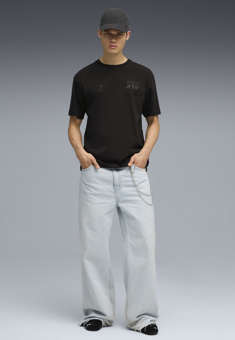 Svart t-shirt med korta ärmar som har grafiska detaljer, kombinerad med ljusblå vida jeans och en svart keps, kompletterad med en kedjedetalj.