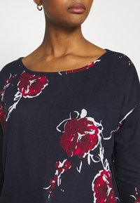 Mörkblå tröja med rundad halsringning, prydd med vita och djupröda blommönster. Tillverkad av ett mjukt, texturerat tyg.