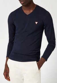 Marineblauwe T-shirt met lange mouwen en V-hals, gemaakt van zachte stof, met een klein rood driehoekig logo op de borst. Minimalistisch ontwerp en aansluitende pasvorm.