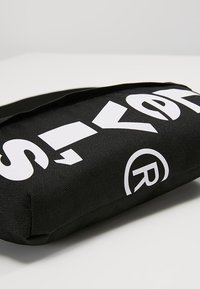 Pochette en tissu noir avec un grand texte blanc en gras partiellement visible, comportant un symbole de marque déposée sur une surface blanche unie.