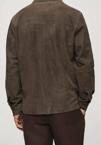 Uomo in giacca scamosciata marrone scuro e pantaloni abbinati, in piedi con una mano in tasca, girato di spalle rispetto allo spettatore.