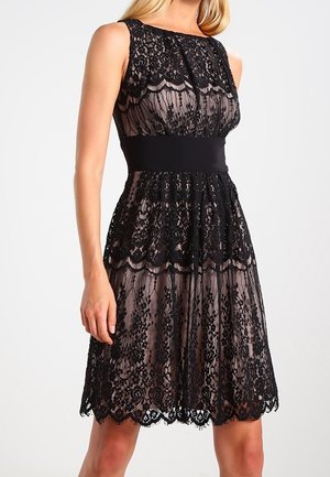 Ärmelloses, knielanges schwarzes Spitzenkleid mit Blumenmustern und einem einfarbigen schwarzen Taillenband, getragen von einer stehenden Frau.