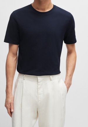 Mężczyzna w teksturowanej granatowej koszuli z krótkim rękawem, włożonej do białych spodni z wysokim stanem i plisami, stoi z jedną ręką w kieszeni.