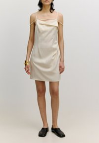 Cremefarbenes Mini-Kleid mit dünnen Trägern, drapierter Ausschnitt und einer leicht strukturierten Silhouette. Getragen mit flachen schwarzen Schuhen.