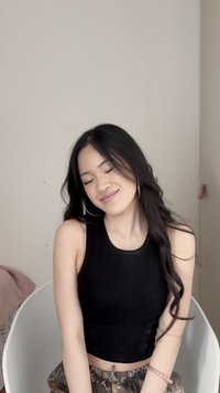 Nuori nainen, jolla on pitkät tummat hiukset ja päällään musta hihaton paita sekä kuvioidut housut, hymyilee lempeästi silmät suljettuina istuessaan valkoisella tuolilla.