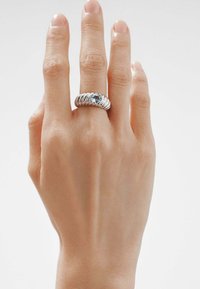 Anillo de plata con diseño retorcido y una piedra central azul, llevado en una mano que muestra dedos ligeramente cuidados contra un fondo neutro.