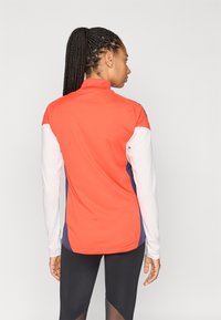 Piros-fehér cipzáras sportdzseki állónyakú, testhez simuló szabással, kontrasztos tengerészkék részletekkel az oldalakon és az alján.