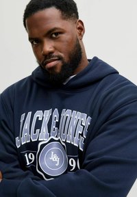 Navy blue hoodie van zacht materiaal. Voorzien van witte tekst "JACK & JONES" en een logo ontwerp, met een groot grafisch detail aan de voorkant.