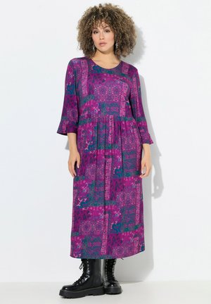 Robe maxi violette avec un design en patchwork présentant des motifs floraux et géométriques. Manches trois-quarts, coupe décontractée, associée à des bottes de combat noires.