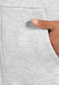 Ruka vkládající palec do přední kapsy světle šedého měkkého oděvu, zobrazení textury látky a detailů šití.