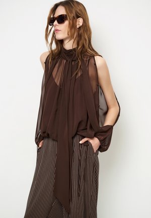 Vrouw met lichtbruin haar die een donkerbruine doorschijnende blouse draagt met een lange sjaalknoop en bruine wijde broek met krijtstreep, handen in de zakken en donkere zonnebril.