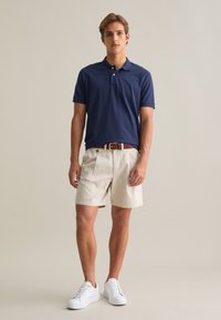 Camiseta tipo polo navy, pantalones cortos beige, cinturón marrón y zapatillas blancas. Mangas cortas, textura de algodón suave, silueta casual y acentos de botones.