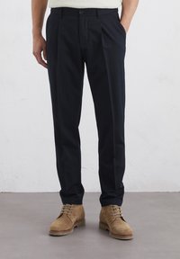 Pantalons bleu marine avec des plis, fabriqués en tissu lisse, associés à des chaussures en daim marron clair avec des lacets et une semelle texturée.