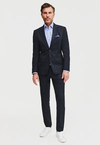 Granatowy garnitur z klapami, połączony z jasnoniebieską koszulą w paski, białymi trampkami i niebieską chusteczką w kieszeni. Dopasowany krój.