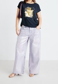 Black t-shirt with gold abstract design, paired with light purple wide-leg jeans. Model wears beige sandals and holds a maroon handbag.