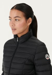 Giacca puffer nera con colletto alto, design a trapuntatura orizzontale e chiusura con zip; presenta una toppa circolare con logo sulla manica sinistra.