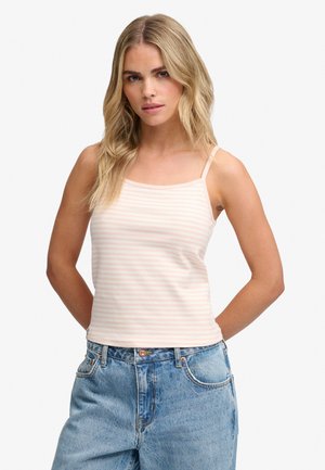 Jeune femme aux longs cheveux blonds portant un débardeur rose clair rayé et un jean bleu ample, debout les mains dans le dos.
