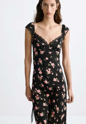 Joven mujer con un vestido negro floral con rosas rosas y ribete de encaje, de pie frente a un fondo liso y claro.