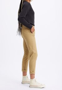 Chinos kaki avec des ourlets retroussés, poches latérales et une texture lisse, associés à des baskets montantes blanches et un sweat-shirt à manches longues foncé.