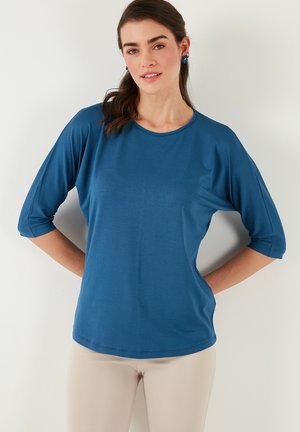 Modrá, volná blůza s kulatým výstřihem a tříčtvrtečními rukávy. Hladká textura a jednoduchý design, vhodné pro neformální nošení.