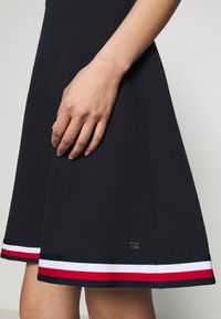 Marine jurk gemaakt van een gladde stof, met een uitlopende vorm. Zoom accentueert met rode, witte en marineblauwe strepen, en een klein metallic logo detail.