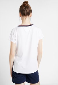 Biała koszulka z krótkim rękawem o okrągłym dekolcie, z granatowym i czerwonym paskiem na kołnierzyku. Gładki bawełniany materiał, luźny krój.