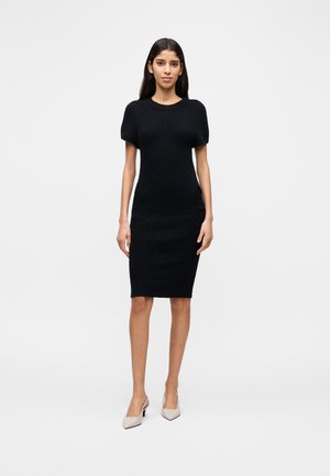 Czarna dzianinowa sukienka z krótkim rękawem, dopasowany fason, teksturowana tkanina, długość do kolan, w połączeniu z jasnobeżowymi szpilkami.