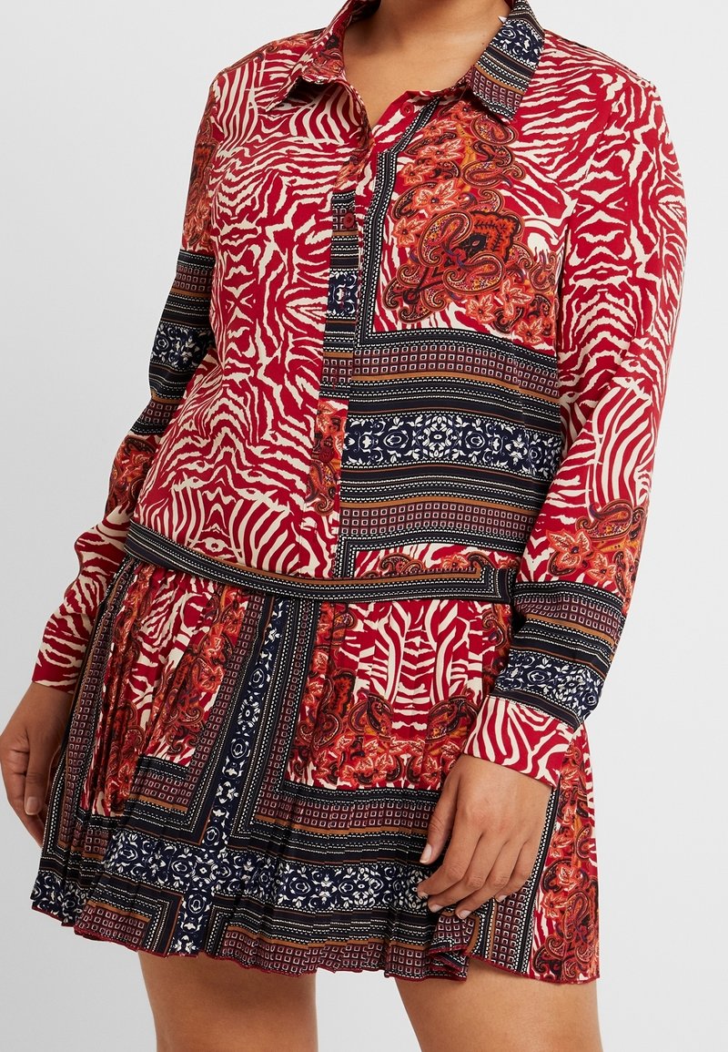 Una modelo lleva un vestido rojo vibrante con patrones llamativos, incluyendo rayas y paisley, con botones y una cintura ajustada, sobre un fondo neutro.