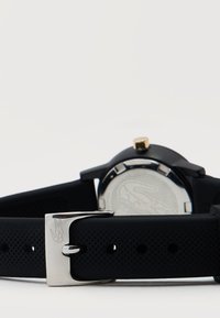 Μαύρο ρολόι χειρός με υφασμένο λουράκι από σιλικόνη, ασημένιο κούμπωμα με χαραγμένο κροκόδειλο και ασημένιο πίσω κάλυμμα με λογότυπο κροκόδειλου.