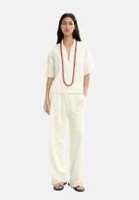 Kremowa bluzka z krótkim rękawem, dekoltem w serek i teksturowanym wzorem, zestawiona z szerokimi spodniami na gumce w pasie. Uzupełniona czerwoną naszyjnikiem z koralików.