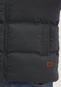 Chaleco puffer negro con cierre de cremallera, que presenta secciones acolchadas, una tela texturizada y un parche de logo de cuero marrón en la parte inferior derecha.