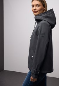 Jeune femme portant une veste à capuche gris foncé avec un col matelassé et un jean bleu, debout de profil contre un mur uni.