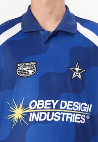 Blue patterned polo shirt with a collar, featuring white accents and embroidered logos: "blizzare NEW VISIONS" and "OBEY DESIGN INDUSTRIES."
