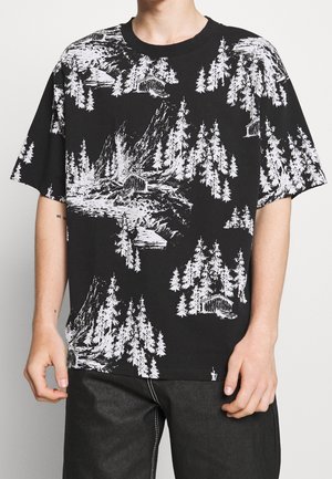 Camiseta negra de manga corta con estampado blanco de árboles del bosque, montañas y cabañas, llevada por una persona con pantalones oscuros.