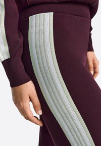 Burgundske športne hlače z teksturirano tkanino in svetlo zelenimi ter belimi navpičnimi črtami na straneh. Prikaz bližnjice z roko na tkanini.