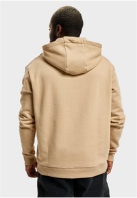 Beige hettegenser laget av mykt stoff, med avslappet passform, ribbede mansjetter og kengurulomme, sett bakfra.