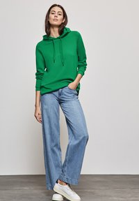 Žena stojící s jednou rukou v kapse, oblečená v zelené mikině, světle modrých širokých džínách a bílých teniskách na šedé podlaze.