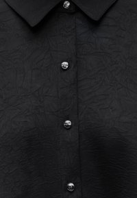 Chemise noire texturée avec des boutons métalliques brillants et un col pointu, montrant un gros plan de la patte de boutonnage avant.