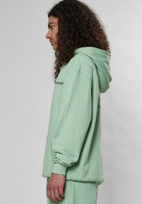 Mintgrön hoodie med en framlapp, dragsko på huvan och svart text på bröstet. Mjuk tyg och avslappnad passform, med ribbade ärmslut.