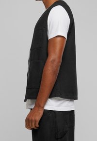 Czarna bezrękawnik z kieszenią z przodu, wykonany z gładkiego materiału, noszony na białej koszulce, charakteryzujący się luźnym krojem i minimalnym wzornictwem.