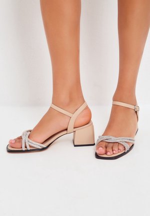 Voeten die beige sandalen met blokhak dragen, met met kristallen versierde kruisriemen en enkelgespen, op een witte achtergrond.