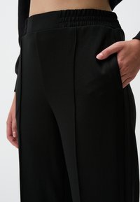 Czarne spodnie wide-leg z elastycznym pasem. Materiał wydaje się miękki i gładki, widoczne są boczne kieszenie.